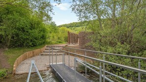 Een houten brug over een beek, met metalen leuningen, omgeven door groen en bomen.