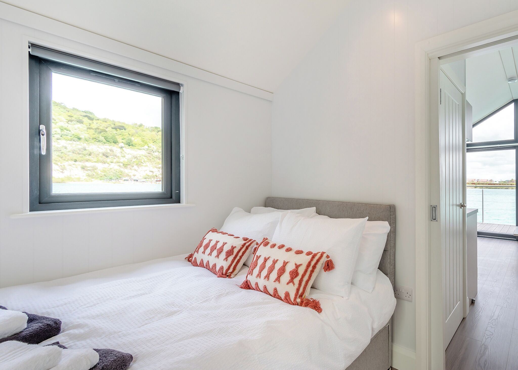 A bedroom with a bed, pillows, a window, and a view of a body of water.