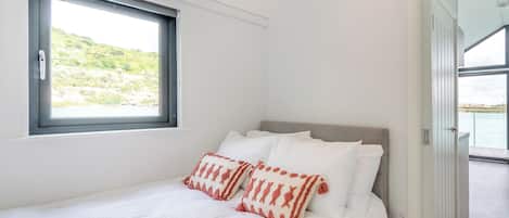 A bedroom with a bed, pillows, a window, and a view of a body of water.