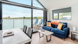 Een moderne kamer met een groot raam met uitzicht op een waterpartij, een blauwe bank, een eettafel en twee blauwe stoelen.