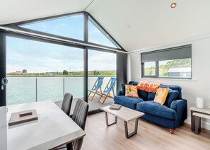 A modern room with a large window overlooking a body of water, a blue sofa, a dining table, and two blue chairs. - 2 bedroom accommodation in Halling (Halling)