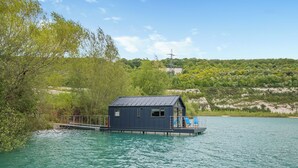 Een drijvend huis met een terras en een klein raam, omgeven door water en groen.
