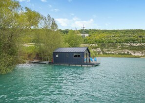 A floating house with a deck and a small window, surrounded by water and greenery.