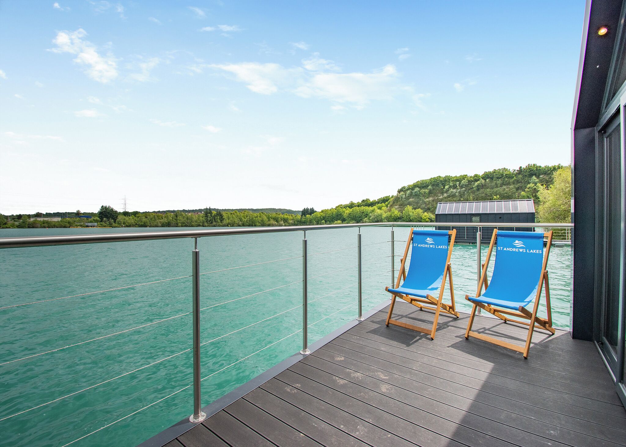 A deck with two blue lounge chairs, a railing, and a view of a calm body of water.