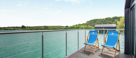 A deck with two blue lounge chairs, a railing, and a view of a calm body of water.