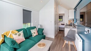 A compact living space with a green sofa, a small dining table, a kitchenette, and a bed in the background.