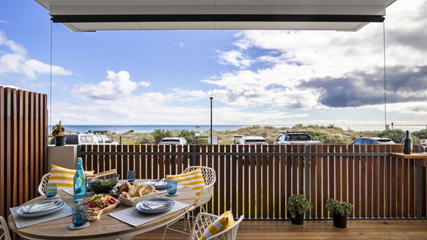 A wooden deck with a table set for a meal, overlooking a beach with parked cars and a cloudy sky. - Moana Beach, Seafront Apartment (Moana)