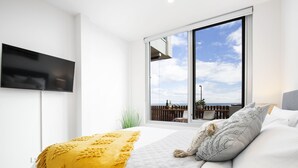A bedroom with a large window, a bed with a yellow throw, and a flat-screen TV on the wall. - Moana Beach, Seafront Apartment (Moana)