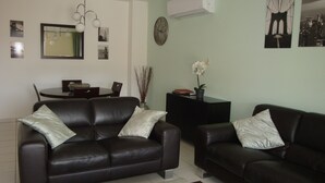 A living room with two black leather sofas, a dining table with chairs, a clock on the wall, and a potted plant.