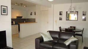 A living room with a brown leather sofa, a dining table with chairs, and a kitchen area in the background.
