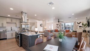 Une cuisine moderne équipée d’appareils électroménagers en acier inoxydable, un coin repas avec une table sombre et des chaises en cuir, et un salon avec un canapé et une grande fenêtre.