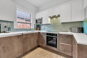 A modern kitchen with white cabinets, a built-in microwave, a coffee maker, and a window with a view of a brick building. - Poets Apartment By My Getaways (Worthing)