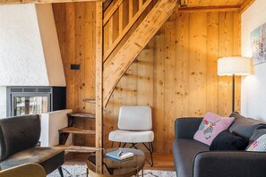 Um quarto acolhedor com uma escada de madeira, uma poltrona cinza, um sofá preto e uma mesa de centro de vidro com um livro.