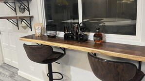Un comptoir de cuisine avec une machine à café, une bouteille et un bloc à couteaux. On y trouve deux tabourets de bar et une fenêtre donnant sur une piscine.