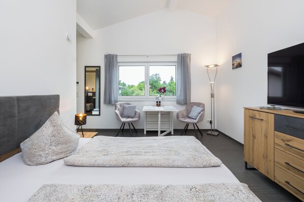 A modern bedroom with a bed, a desk, two chairs, a TV, and a window with curtains. - 25-square-meter apartment with a balcony and a separate bedroom (Bad Feilnbach)