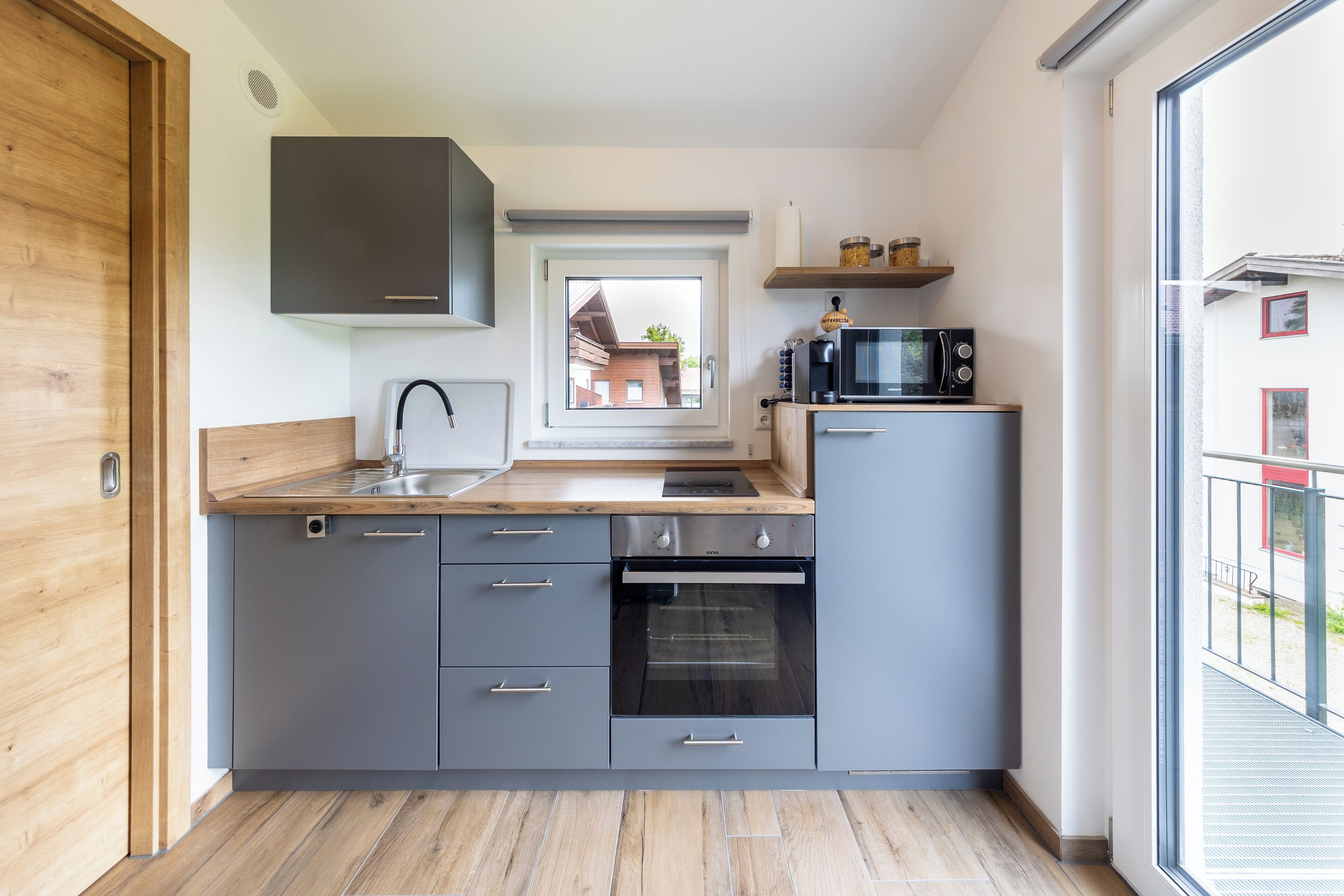 Una cocina moderna con armarios grises, horno empotrado y ventana con vista a un barrio residencial.