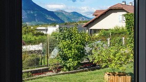 Une vue depuis une fenêtre d’une maison avec un jardin, une montagne en arrière-plan et un ciel dégagé.