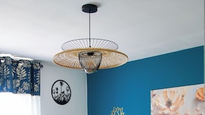 Un lit avec une couette jaune, des oreillers bleus et une décoration florale. On peut voir une œuvre d’art représentant des fleurs accrochée au mur, une horloge murale noire et un luminaire suspendu.