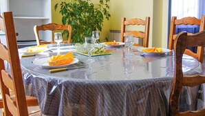 Une table de salle à manger ronde, recouverte d’une nappe transparente, est entourée de chaises en bois. On y voit des assiettes remplies de nourriture, des couverts, et une plante en pot en arrière-plan.