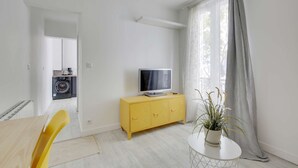 A modern living room with a yellow cabinet, a flat-screen TV, a dining area with a wooden table and yellow chairs, and a kitchen visible through an open door. - Chic studio near Paris (Colombes)