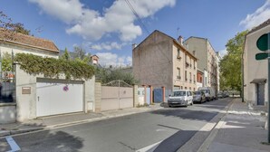 A street with parked cars, a gated entrance, and buildings with balconies. - Chic studio near Paris (Colombes)