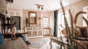Ein gemütliches Wohnzimmer mit einem Holztisch, einer Couch mit gemusterten Kissen, einem großen Spiegel über einem Holzschrank und einem Fenster mit Vorhängen.