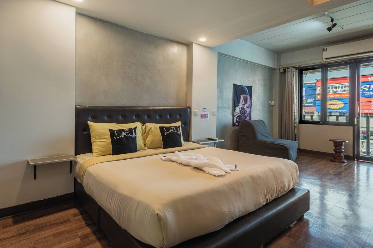 Quarto de hotel com uma cama grande, uma mesa de cabeceira, uma cadeira e uma janela com vista para contentores de transporte.