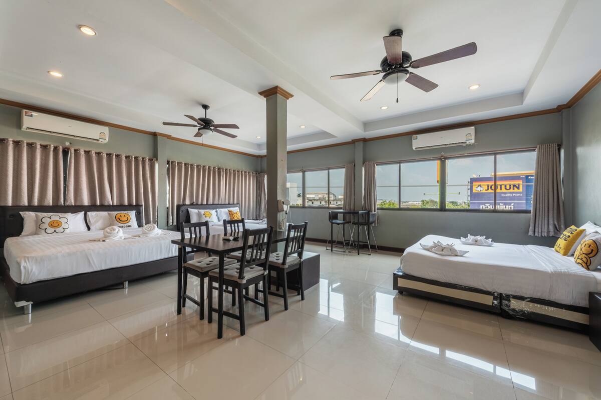 Quarto de hotel com duas camas, uma mesa de jantar, cadeiras, ventiladores de teto, janelas amplas e aparelhos de ar condicionado.