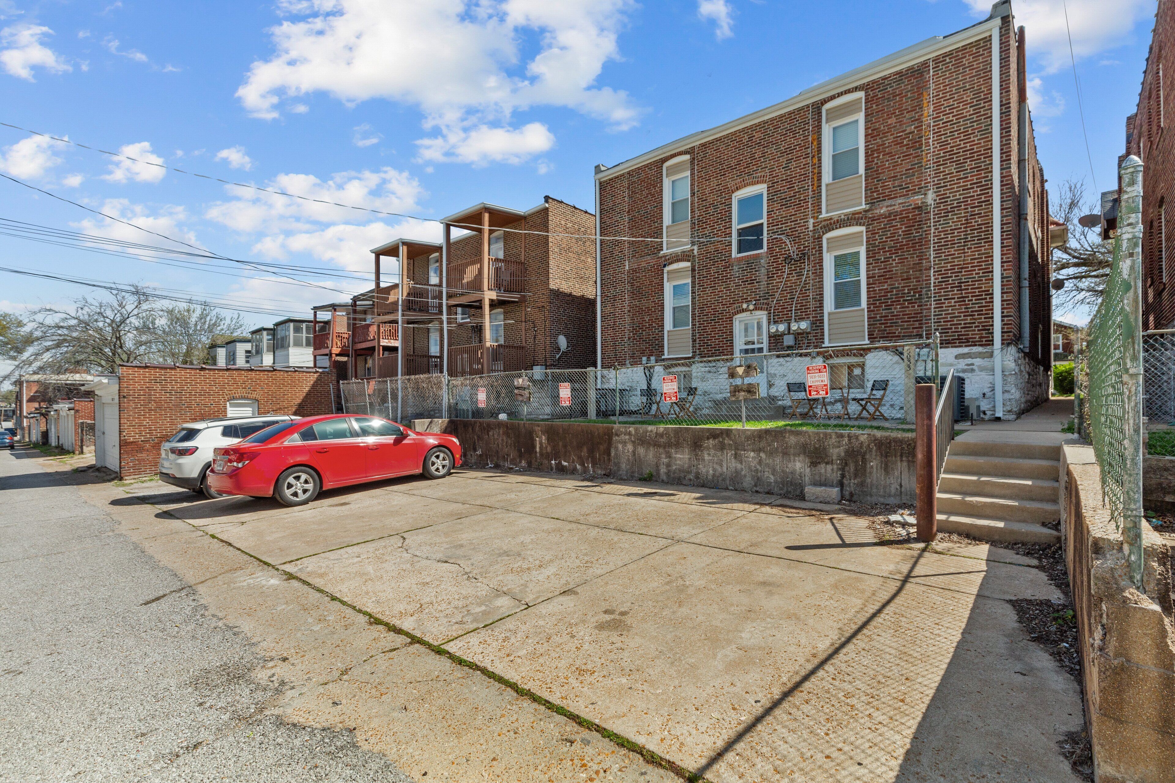 A parking area with a red car and a white car parked. There is a brick building with multiple windows and a balcony.