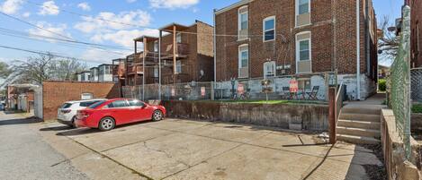 A parking area with a red car and a white car parked. There is a brick building with multiple windows and a balcony.