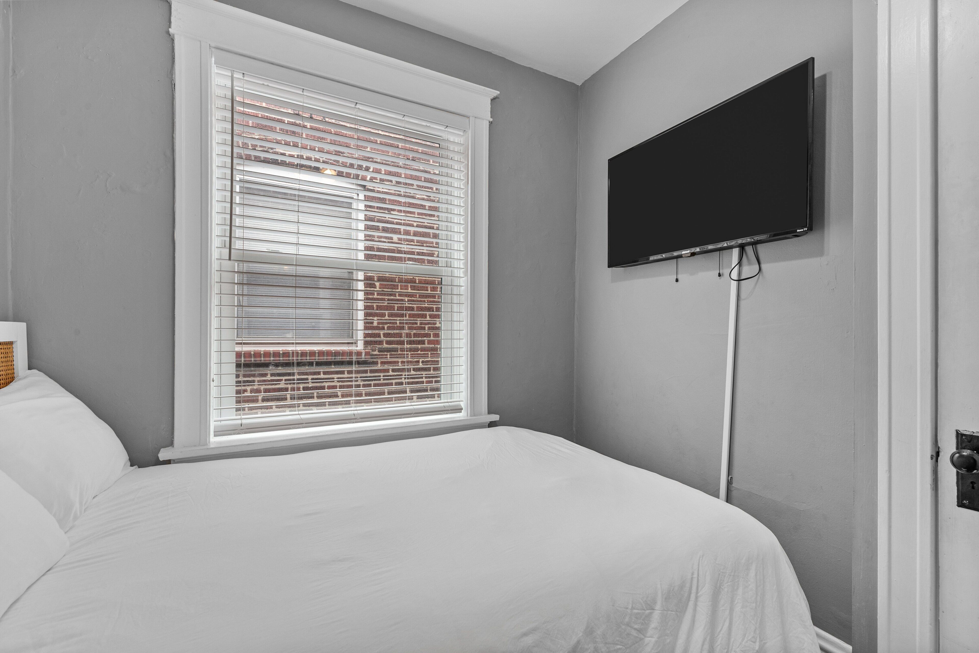 This reverse second-bedroom angle shows the wall-mounted TV, white-dressed bed, large window, gray walls, white-trimmed door, and hardwood flooring.