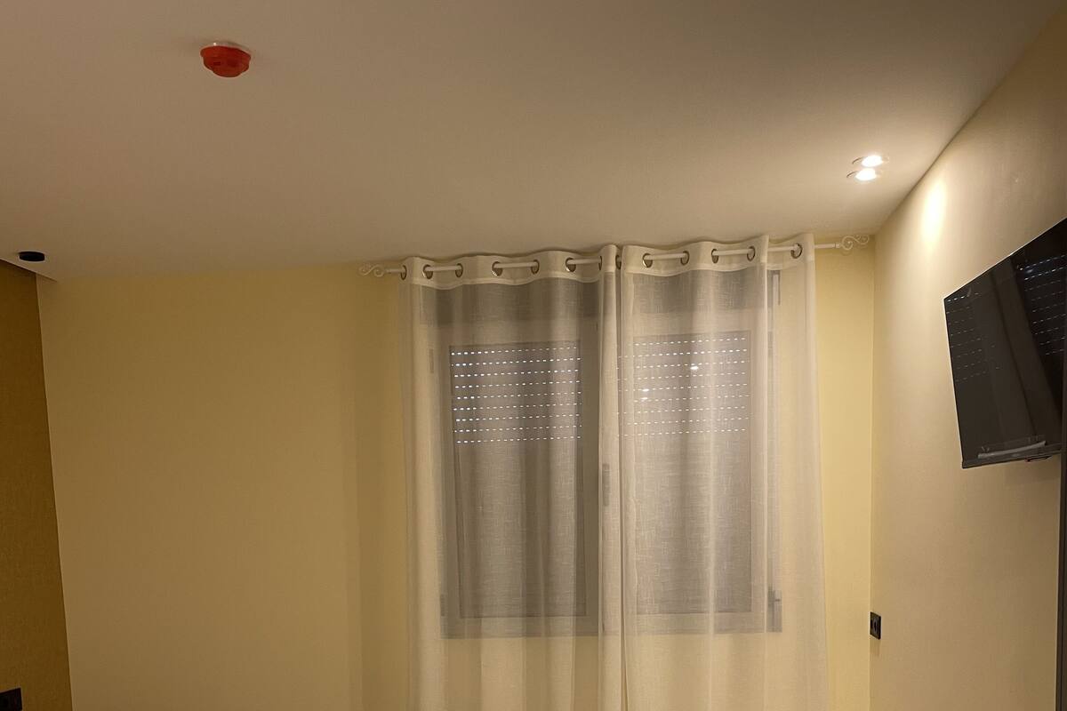 A hotel room with two beds, a desk, a chair, a TV, and a window with sheer curtains.