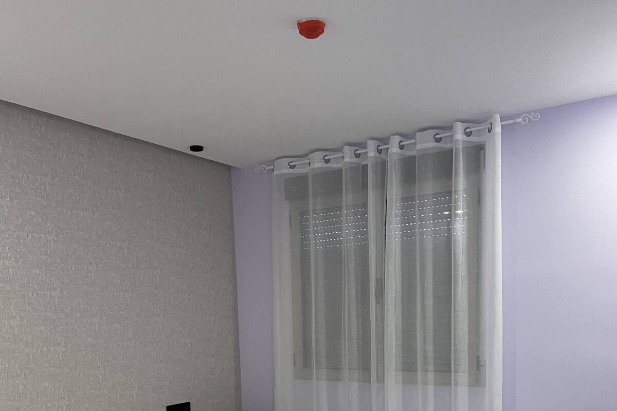 A bedroom with a bed, a nightstand, a desk, and a window with sheer curtains.