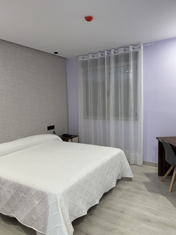 Un dormitorio con cama, mesita de noche, escritorio y ventana con cortinas traslúcidas.