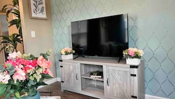 Un salon avec un téléviseur à écran plat posé sur un meuble en bois, deux compositions florales et une grande plante en pot.