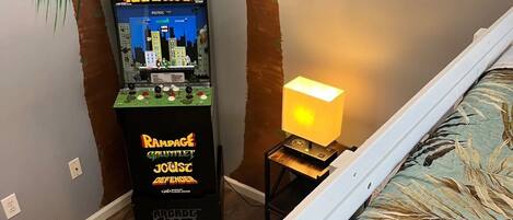 Een slaapkamer met een bed, een nachtkastje met een lamp, een wandgemonteerd spelcomputer en een grote kunstpalm.