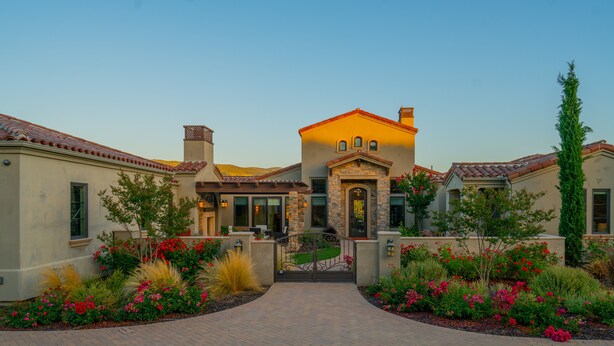 A two-story house with a central entrance, surrounded by a landscaped garden with red flowers and shrubs. - Mansion on De Portola with Vineyard and Mountain view (Temecula)
