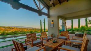A patio with wooden furniture, a hanging chair, and a view of a landscaped garden and distant mountains. - Mansion on De Portola with Vineyard and Mountain view (Temecula)