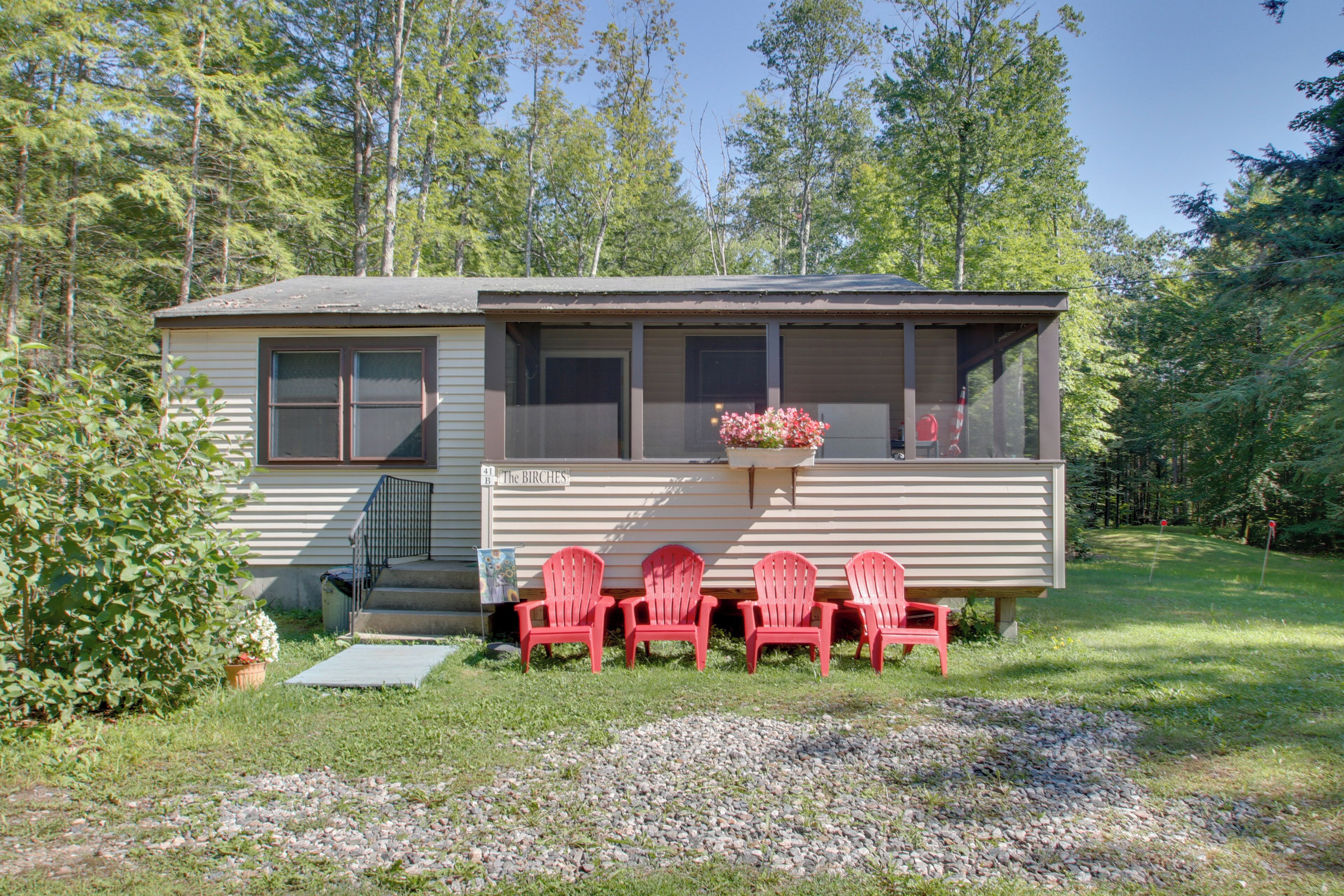 A small cabin with a porch, four red Adirondack chairs, and a flower pot on the window sill.