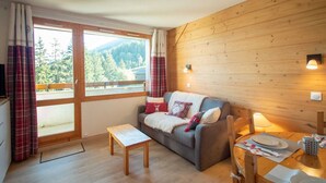 A cozy room with a wooden wall, a grey sofa with red and white cushions, a wooden table with plates and a mug, and a window with curtains.