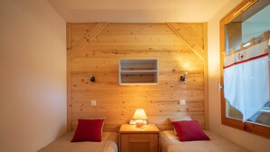 A small, cozy room with two beds, a nightstand with a lamp, a shelf above the beds, and a window with a curtain.
