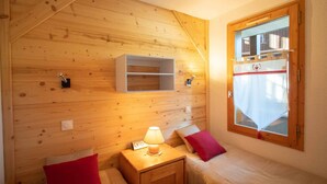 A cozy room with wooden walls, a small bedside table with a lamp, two beds with red pillows, and a window with a view of a building.
