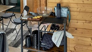 Un área de cocina con una mesa de madera, sillas, un carro metálico negro con utensilios de cocina, un tazón de vidrio transparente y un hervidor.