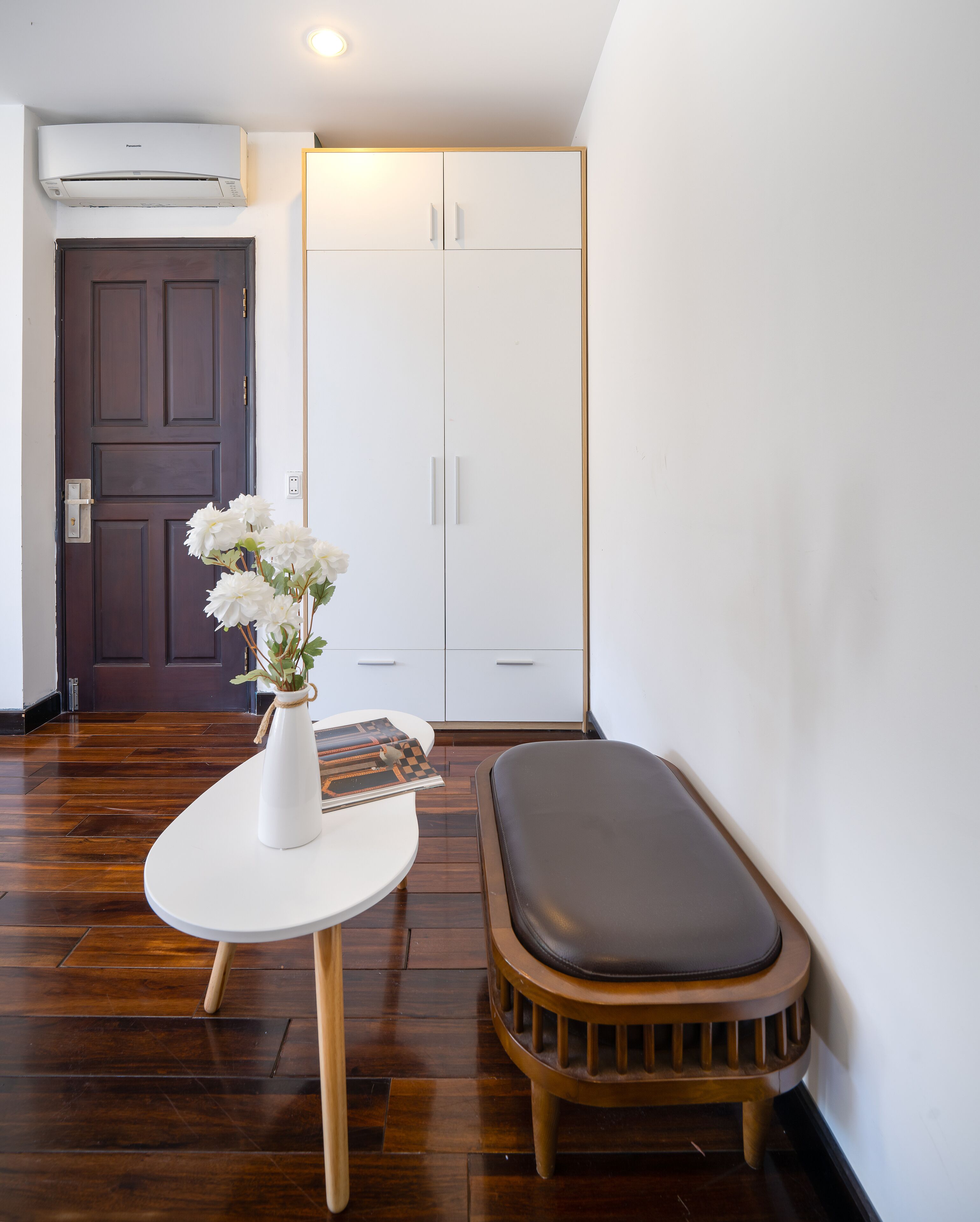 A room with a white wardrobe, a dark wooden door, a white vase with flowers, a small white table, and a wooden bench.