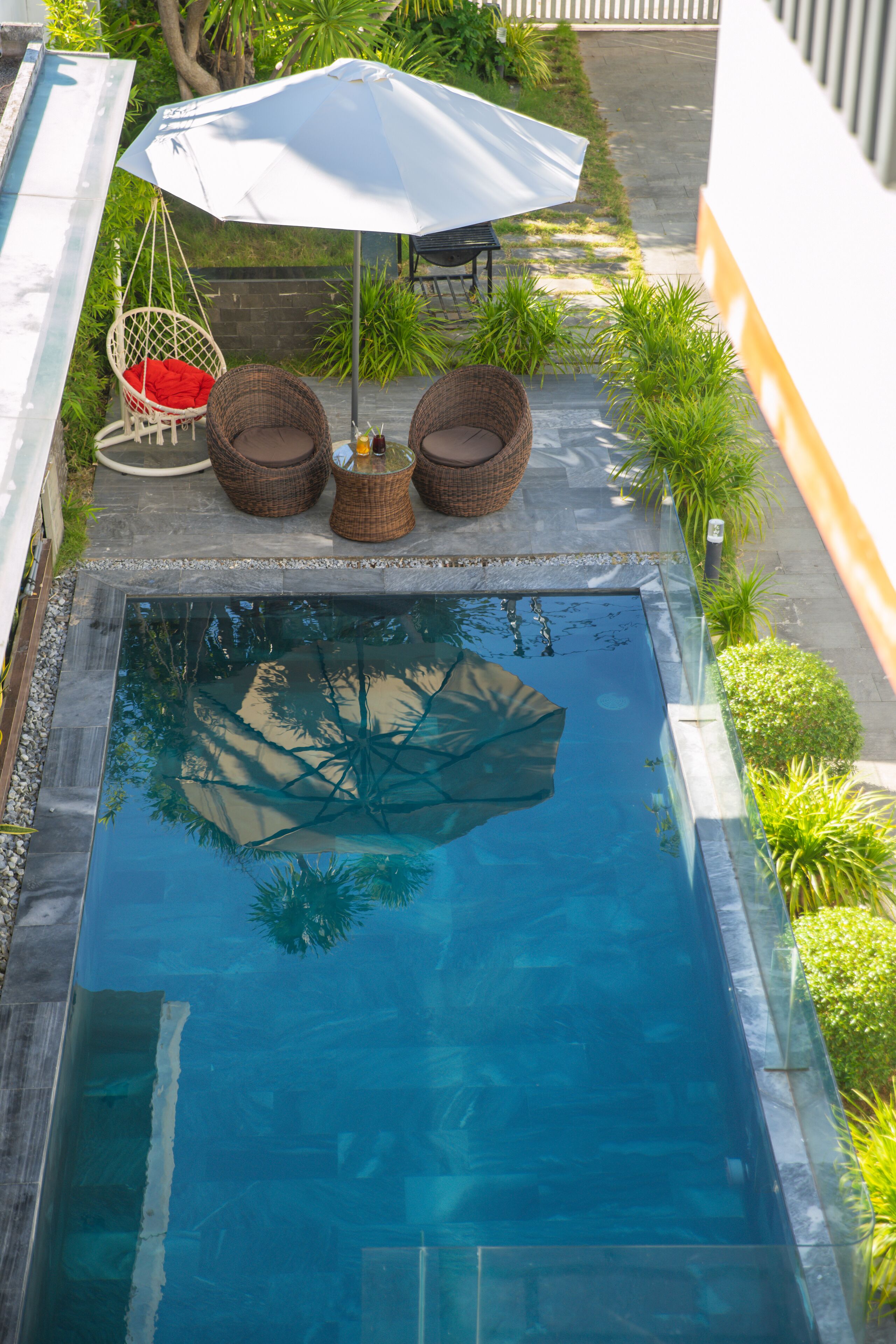 Una piscina rectangular amb superfície d'aigua blava i cristal·lina, envoltada per una tanca de pedra. Hi ha cadires de vimet i una cadira penjant sota un parasol blanc, i petites plantes verdes al voltant de la piscina.