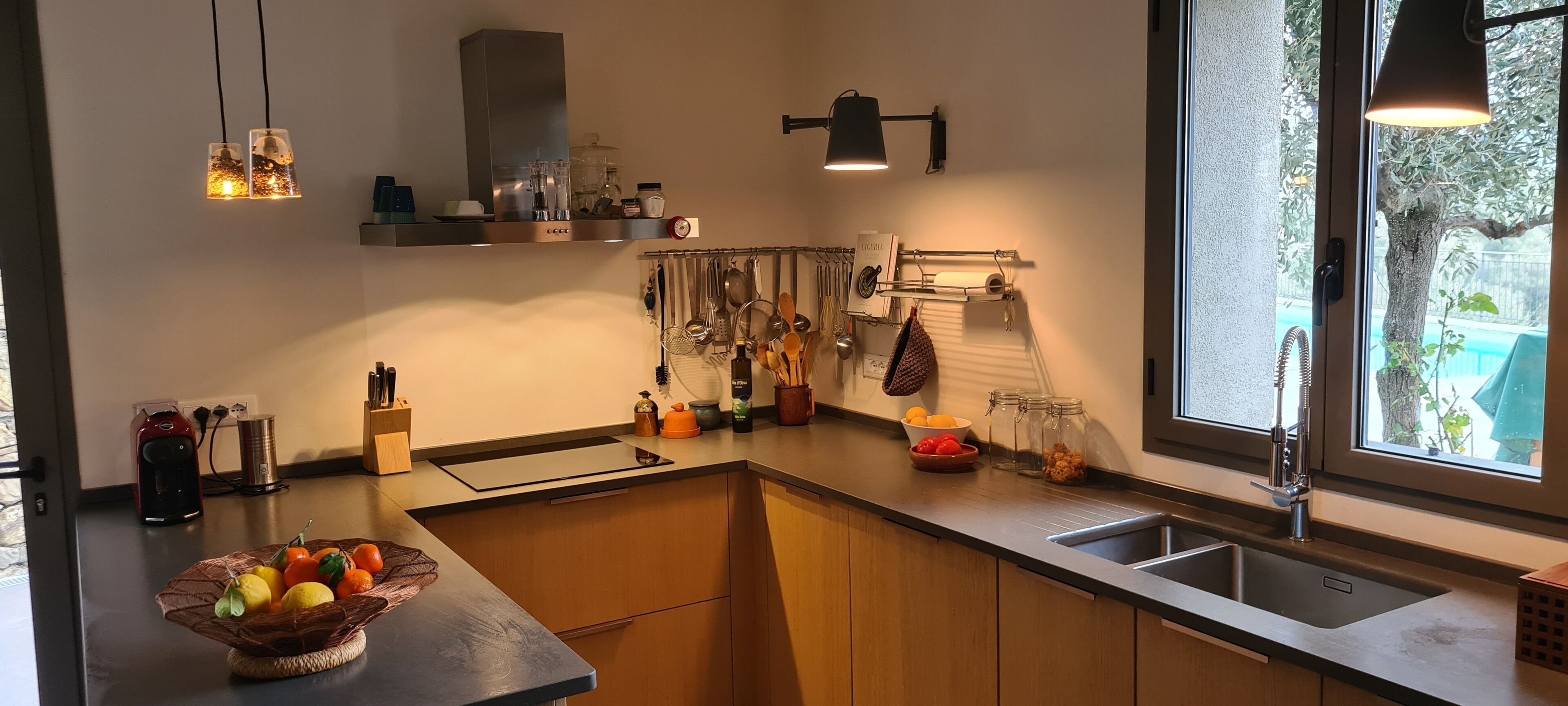A modern kitchen with wooden cabinets, a stainless steel sink, hanging utensils, and a fruit bowl on the counter.