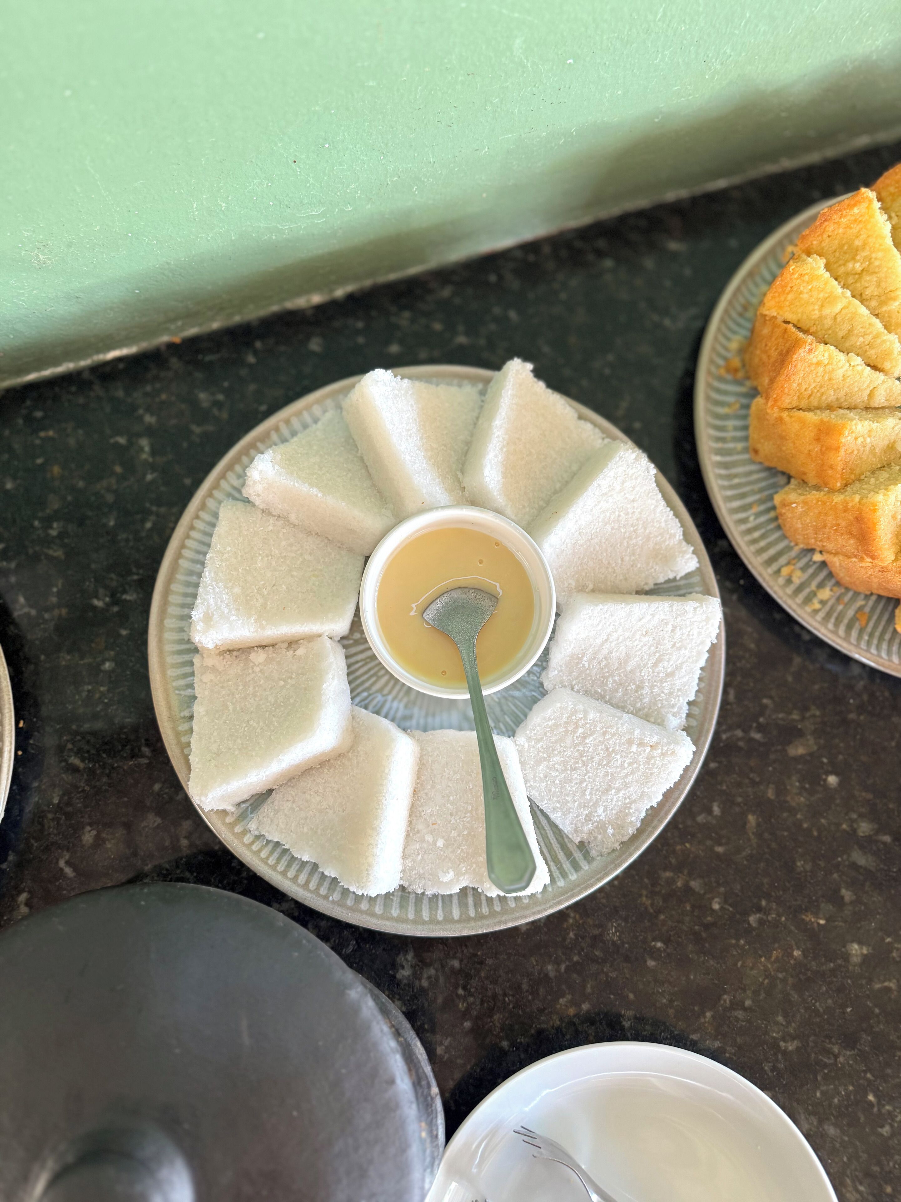 A plate of white, sugar-coated items with a spoon, a plate of golden-brown baked goods, and a metal container on a dark countertop.