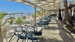 Un patio frente a la playa con mesas y sillas, una pérgola y vista al océano.