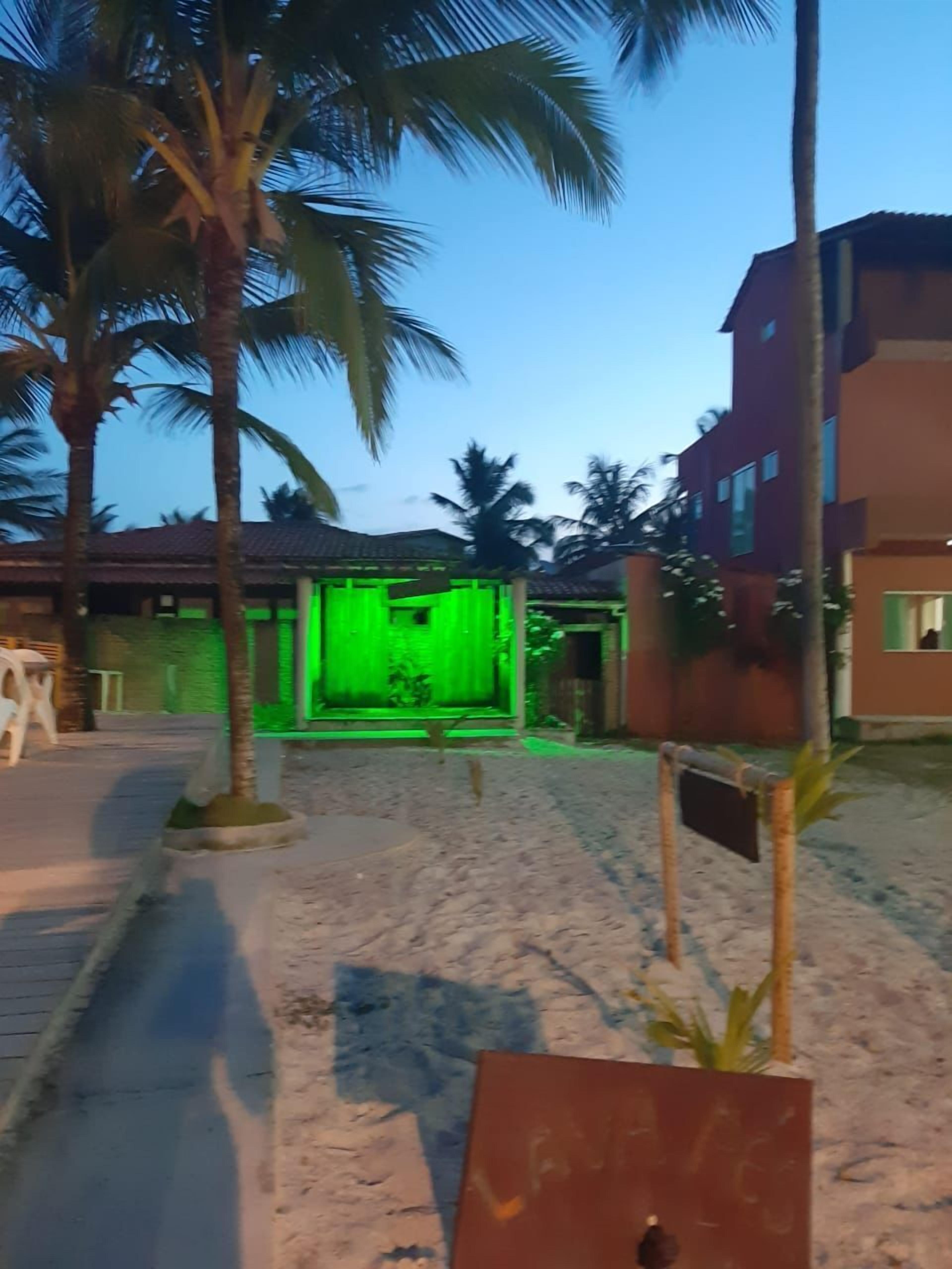 Un sentiero sabbioso conduce a un edificio con interni illuminati di verde, circondato da palme e un edificio rosso sullo sfondo.