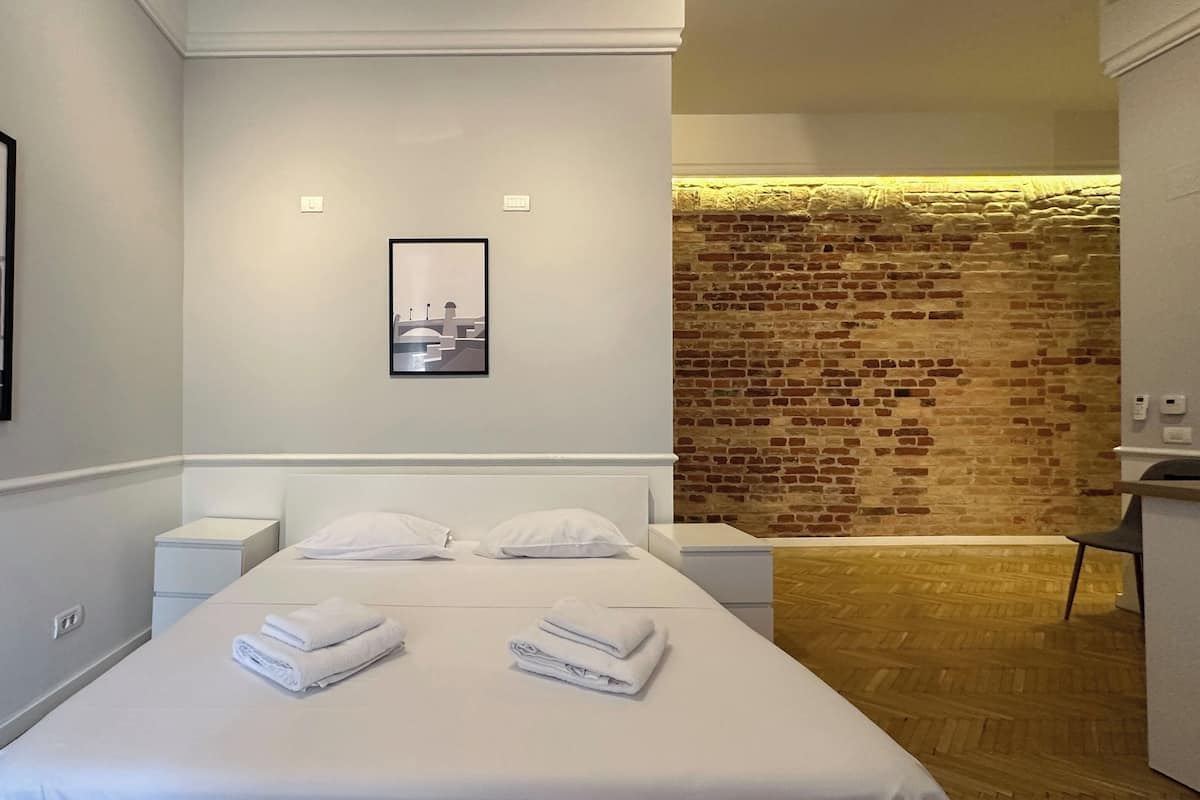 Habitación de hotel con una cama, dos mesitas de noche, un cuadro enmarcado en la pared y un muro de ladrillo al fondo.